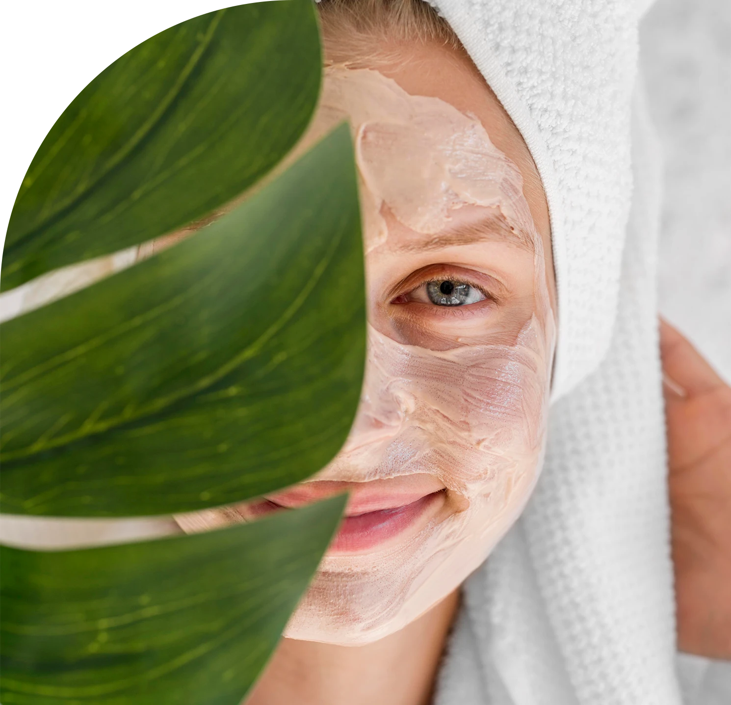 Eine Frau hat eine Gesichtsmaske im Gesicht und trägt einen Handtuchturban. Sie schaut zufrieden in die Kamera. Die Hälfte ihres Gesichts wird mit einer Pflanze bedeckt.